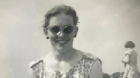 Margaret Raymond A black and white photo of a young woman wearing sunglasses and a summer dress, with another woman in the background facing away from the camera.