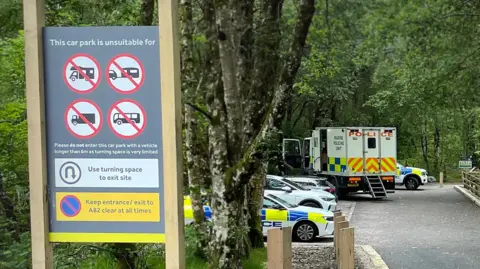 BBC Police cars and an ambulance parked in a car park in the background surrounded by trees. A sign in the foreground signals that the car park is unsuitable for certain large vehicles like trailers, lorries and buses 