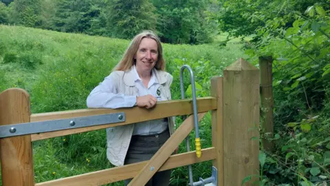 LDRS Deborah Fox, chief executive of the Malvern Hills Trust, has long blonde hair and is wearing a white shirt, dark skirt and a beige waistcoat. She is standing behind a wooden gate on the hills. There is a field behind her and shrubs and trees around her.