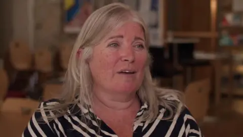 BBC Professor Mackay has shoulder-length blonde hair and is wearing a shirt with diagonal black and cream stripes. The room behind her is blurred with a bookcase visible