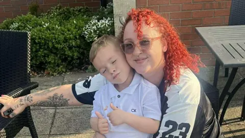 A woman woth red curly hair and sunglasses is crouched down and hugging a young by in a white polo shirt. The boy has his thumbs up and they appear to be in a garden 