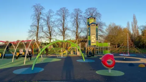 An image of a large green play area, with a large metal slide and netted bridge