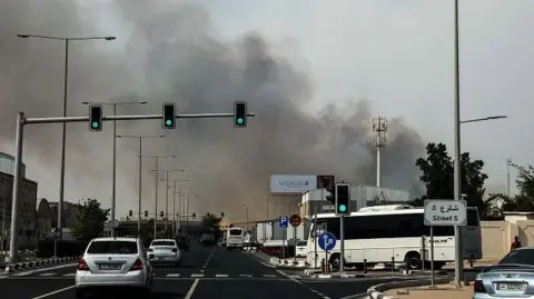 Getty Images Motorists drive past a plume of smoke rising from a reported Iranian strike in the industrial district of Doha on 1 March.