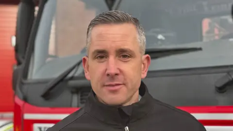 George King/BBC A head and shoulders image of Henry Griffin. He is wearing a black jacket and standing in front of a fire engine. He is looking into the camera. 