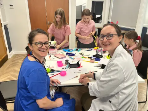 Two women smile at the camera across a table covered with toy parts, tools and brightly coloured offcuts, while schoolgirls in pink gingham uniforms take apart toys behind them.