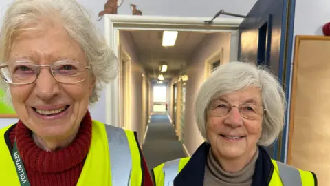 Two older ladies with white hair, wearing hi-viz and a corridor is behind them with rooms off.