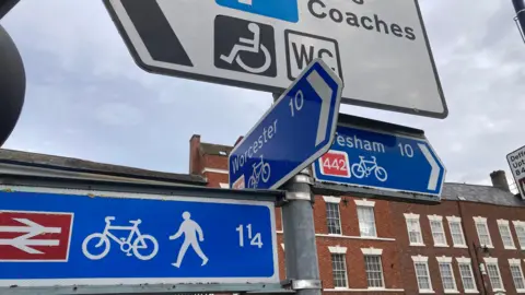 A street sign, showing a left turn towards Worcester and a right turn towards Evesham. There are some buildings in the background. 
