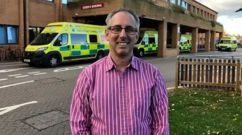 A man in a pink striped shirt stands in front of a hospital emergency department . Behind him are several ambulances. 