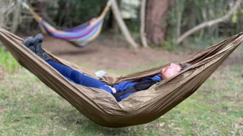 A man with a beard and wearing a blue jacket and trousers is lying in a brown hammock with his eyes closed. Behind him you can see another colourful hammock among trees.