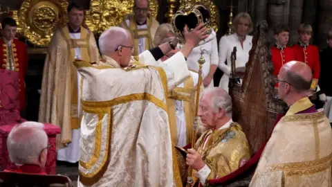 O arcebispo da Reuters, Justin Welby, segura uma coroa sobre a cabeça do rei Charles enquanto Charles se senta no trono. Ambos usam vestes douradas.