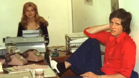 Piccadilly Radio Tony sits with his leg up on the table wearing a bright red shirt and pinstripe navy suit trousers. There are lots of telephones and typewriters around him. There is a woman to the left of the picture with mousy brown hair and a black top on. 