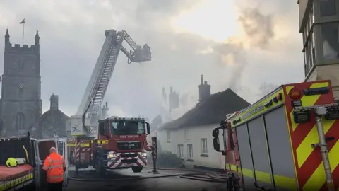 Multiple fire crews are pictured in the village. One fire engine is positioned on support stilts while a ladder extends from the rear of the vehicle so firefighters can squirt water over the thatched-roofed cottages. 