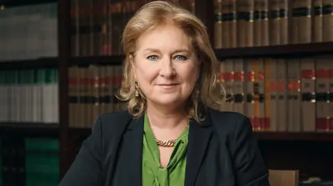 Rory Lindsay/UK Judiciary Baroness Sue Carr sits in front of a bookshelf with her arms crossed. She's wearing a green shirt and black suit jacket. 