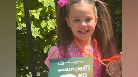 Claire McFaul A young girl, Mya pictured at the Dance World Cup. She is wearing a pink dress with rhinestones and a tutu skirt. Her hair is in pigtails with a pink bow clipped onto the left one. She stands for the picture holding a printout saying, "World finals third place" and in her right hand she is holding her medal. 