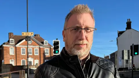 Clare Worden / BBC Ian Milligan looks directly at the camera. He is wearing a black leather jacket, has glasses on and has a white beard. He is standing on a pavement outside a Georgian red-brick building, with 11 sash windows to its front and a blue door. The sky is clear blue. 