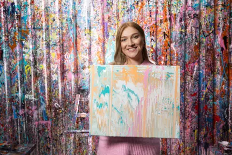 RNID A smiling Caris Lount with brown shoulder-length hair wears a pink jumper. She is holding a multi-coloured piece of splatter-paint art. She is standing in front of a wall also covered in splatter paint. 