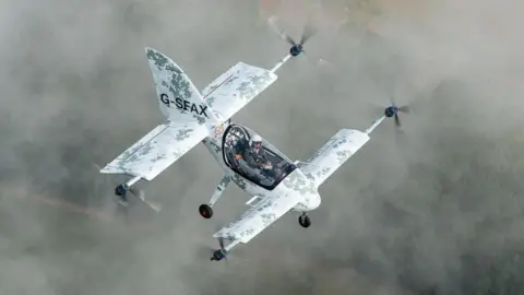 The Axe flying in a misty sky and featuring eight electric motors – two on each wingtip - it has a white and grey camouflage style appearance and Dr William Brooks can be seen sat in the cockpit with his white helmet on his head. 