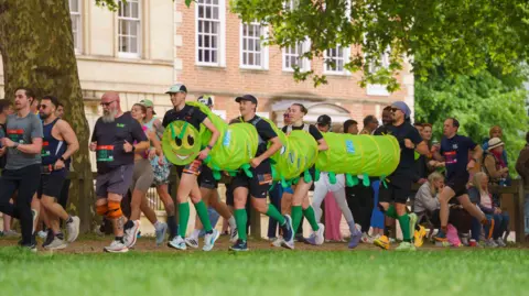 AJ Bell Great Bristol Run Four people running together dressed as a caterpillar. They are running through Queens Square amongst other participants.