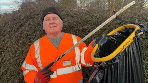 John Devine/BBC A man wearing an orange high-vis jacket and a black woolly hat, holding a litter-picker and a black bin bag.