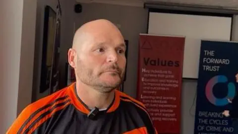 A bald man wearing an orange and black shirt while stood in a small conference room