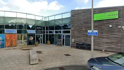 Google Maps A wide image of the exterior of the bistro building which is located next to a bike shop. It is a glass building located behind a carpark. 