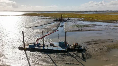 PA Media A shot of the restoration work being done at Lymington. Equipment can be seen set up on mud next to a body of water. 