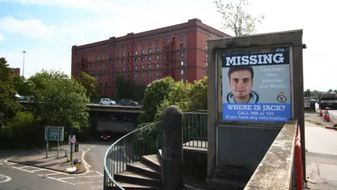 A large missing poster of Jack O'Sullivan posted on a bridge in the area where he was last seen. There is a big red-brick industrial building in the distance.