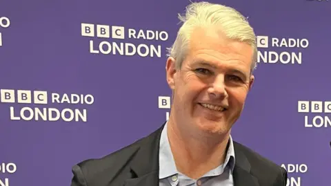 Dominic Twomey is seen from the shoulders up standing in front of a purple backdrop with BBC Radio London signage repeated across it.