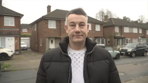 A greying hair man wearing a black puffer jacket and a white shirt. He is standing on a residential road with cars parked behind him.