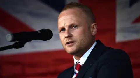 Getty Images Jamie Bryson speaking into a microphone at a 2021 protest against the post-Brexit Irish Sea trade border. He has short blonde hair and is wearing a navy suit and union flag tie. There is a union flag in the background.