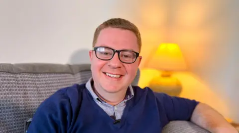 Martin Giles / BBC A smiling sandy haired man with dark glasses. He is wearing a navy blue V-neck sweater over a patterned shirt. He is sitting on a sofa with a lamp in the background. 