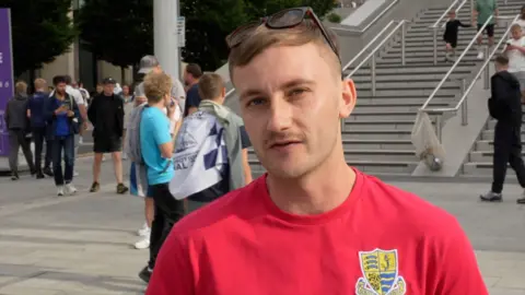 A man wearing a red T-shirt with the Southend United logo on the front while standing outside Wembley Stadium.
