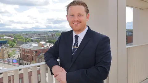 Head-and-shoulders portrait of Chris Eynon. He is wearing a black suit, white shirt and dark tie. He is standing outdoors near a light-colored building. He is smiling. In the background you can buildings and a church beneath a cloudy sky. It is daytime. 