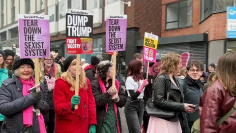 Graeme Maxwell/BBC Women hold placards that read 'tear down the sexist system' and 'dump trump' as they wear bright colours and walk together through the streets of Brighton