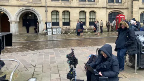 A woman and man in modern padded coats in the foreground with film extras wearing 1940s-style clothing in the background