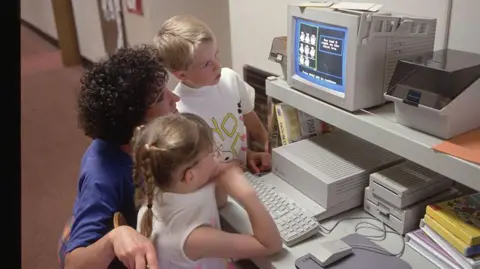 Shows a classroom assistant with dark curly hair and a purple t-shirt kneeling down, talking to a young boy and girl with fair hair and white t-shirts. They are looking at a computer. 