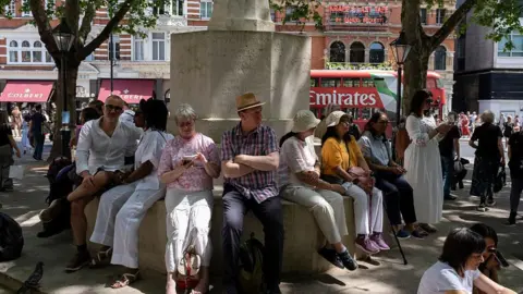 Getty Images A group of people sit on a stone ledge under the shade of trees in central London. 