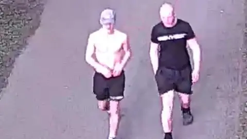 Gloucestershire Police Two men in blurred CCTV footage. One is shirtless, and wears black shorts and a grey cap. The other is bald, and wears a black t-shirt, shorts and trainers. They are on a wide path with grass either side of them. 