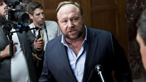 Conspiracy theorist Alex Jones, wearing a blue suit, stands at a microphone in front of news cameras 