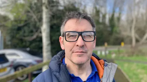 Laura Coffey/BBC A man is standing in front of some grass and parked cars. He has short greying hair, is wearing thick black glasses and wearing a blue polo shirt and navy jacket. 