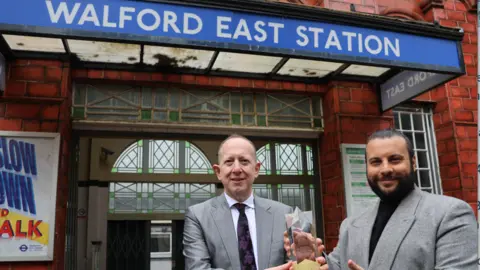 Hertsmere Borough Council Jeremy Newmark is wearing a grey suit and stood near the entrance to Walford East Station as he passes an award to Chris Clenshaw