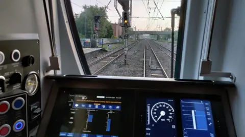 The picture shows the view of a train track through the window of a train driver's cab. Alongside the tracks, a traffic-light style signal can be seen glowing amber. The train driver's electronic dashboard and some dials and buttons can be seen in the foreground.