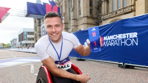 Manchester Marathon Callum Hall smiles as he holds a blue accolade saying "1" in front of the main stone building of the university. A blue banner saying Manchester Marathon is behind him.