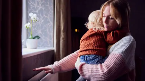 A woman putting her hand over a radiator, while holding her child in her arms. She is wearing a jumper as is the child. A windowsill can be seen in the left of the picture.