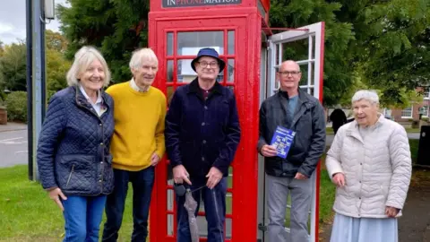 The image shows five people standing in front of a red telephone box. The group consists of three women and two men, all smiling. One man holds a pamphlet, and another holds an umbrella. They are all dressed in casual, weather-appropriate clothing, with jackets and sweaters. Trees and a grassy area are visible in the background.






