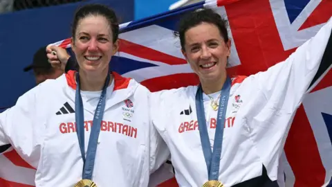 Two women with dark hair wearing white Great Britain Olympic kits. They're both wearing gold medals around their necks, smiling and holding a Union Jack behind them.