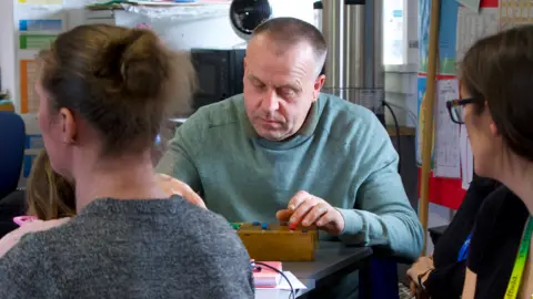 A man sat at a table during one of the University of Sheffield's dementia workshops, held in partnership with charity Age UK. He is sat at a table, focused on a wooden box with audio faders built into it. He's being watched by a woman wearing a university lanyard.