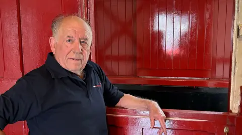 Supplied David McKay is leaning on the red wooden frame of the carriages and looking just beyond the camera. He has short, balding grey hair and is wearing a dark blue shirts with a red logo on the the chest.