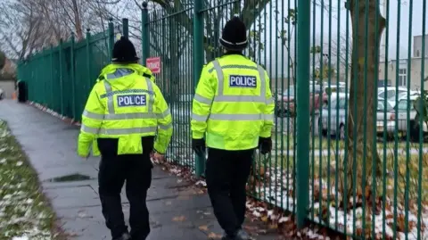 Cleveland Police Two police officers wearing hi-vis jackets walk along a pavement. They are walking past a building behind a large, green fence.
