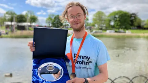 Lewis Adams/BBC Joe Griggs holds up his prize - a silver plate - and is wearing a medal. Behind him is the muddy estuary at high tide.